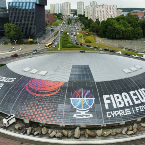 Widok na halę sportową z logo FIBA EuroBasket 2025 na dachu, z otaczającą infrastrukturą drogową.